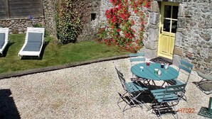 Un edificio in pietra con un patio in ghiaia, un piccolo giardino con fiori rossi e un'area salotto esterna con tavolo e sedie.