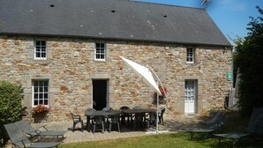 A stone building with a tiled roof, a patio with a table and chairs, and a garden area with lounge chairs. - Semi-detached house with 4 bedrooms at Manoir de la Barguignerie (Saint-Christophe-du-Foc)