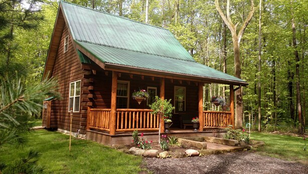 Une cabane en rondins avec un toit en métal vert, une véranda en bois et un petit jardin à l’avant.
