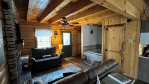 L’intérieur d’une cabane en rondins comprend un salon, une cheminée, un mur en pierre, un ventilateur de plafond et une salle de bain avec une baignoire.