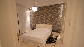 A bedroom with a stone wall, a bed with a white bedspread, a bedside table, and a modern chandelier. - Welcome Home (Sousse)