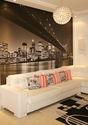 A white sofa with patterned cushions, a black and white cityscape mural on the wall, and a chandelier.