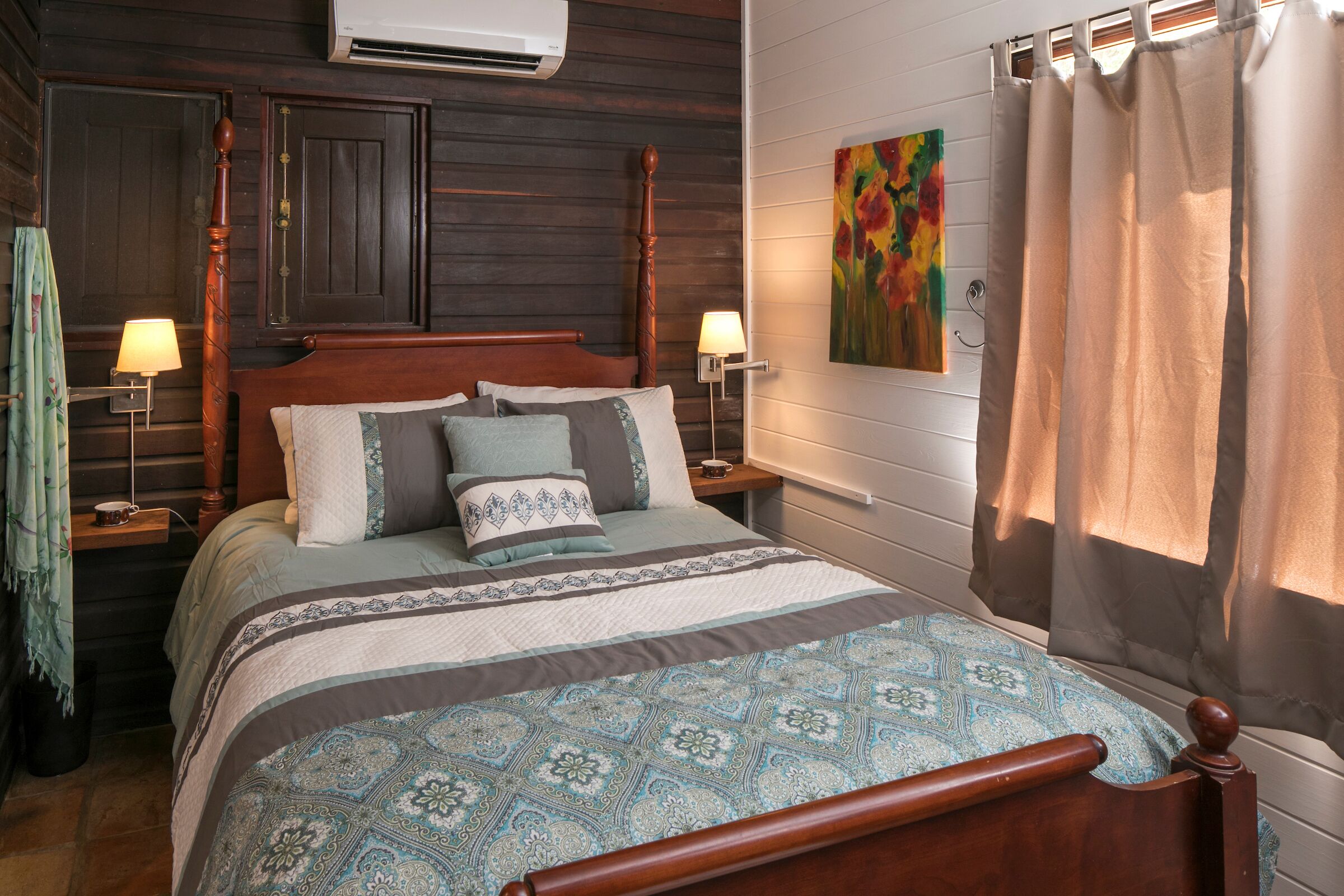 A neatly made bed with a patterned bedspread, two bedside lamps, a wall-mounted air conditioner, and a colorful painting above the bed.