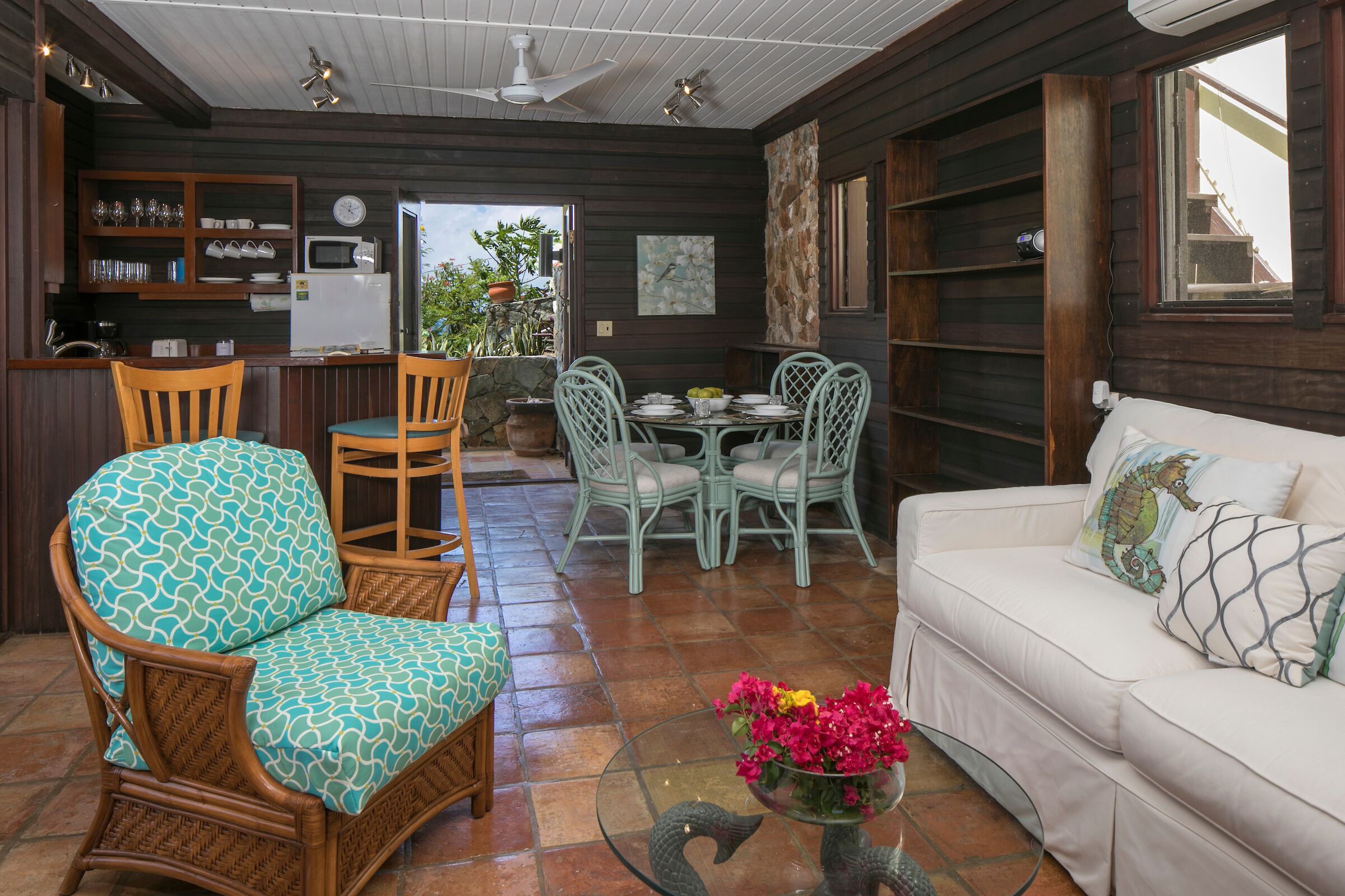 Un espacio de sala acogedor con muebles de mimbre, una mesa de centro de vidrio con flores y un sofá blanco con cojines decorativos.