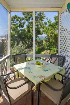 Eine überdachte Terrasse im Freien mit Tisch und Stühlen, einer Obstschale und Blick auf Grünflächen und Gebäude.