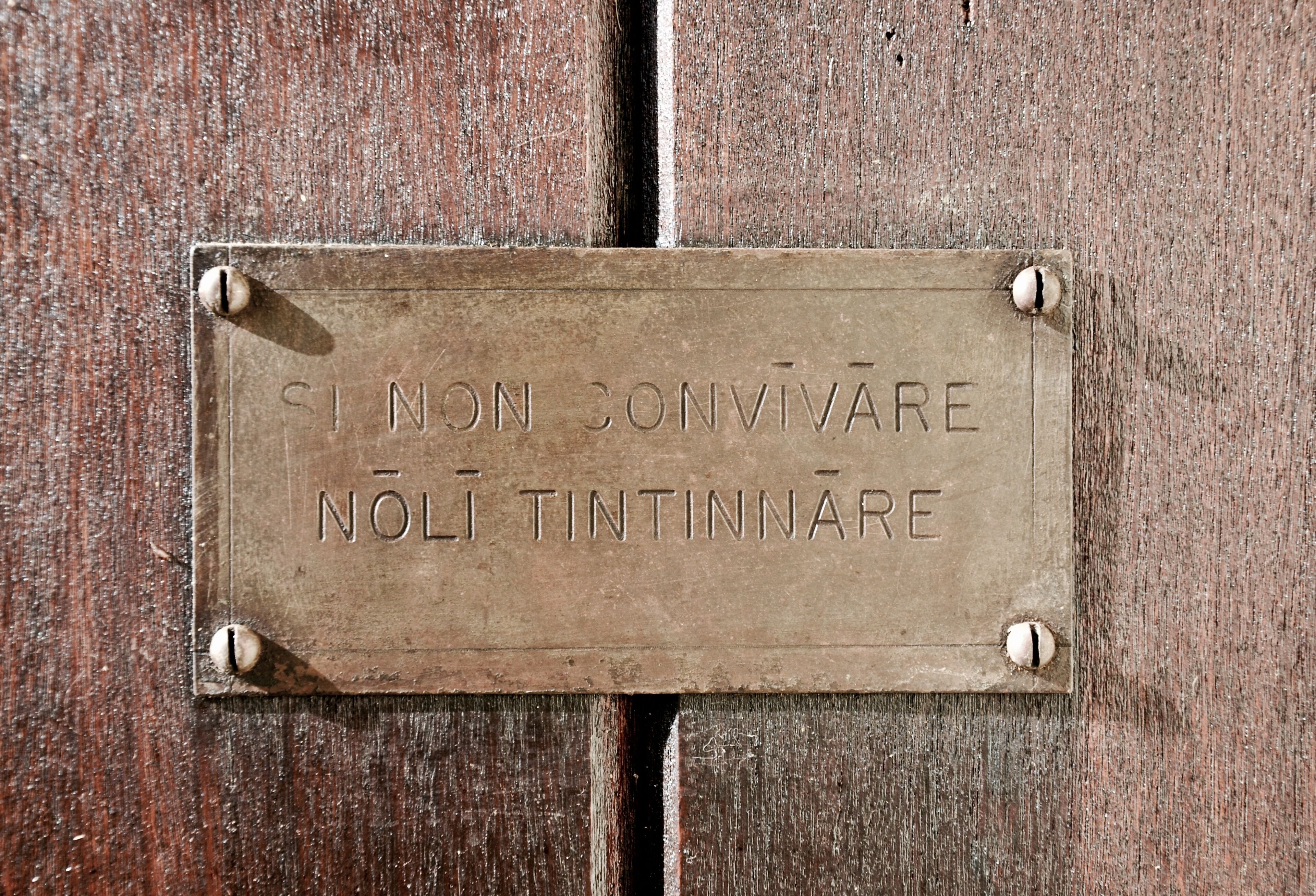 En metaldel med indskriften "SI NON CONVIVARE NOLI TINTINNARE" monteret på en træoverflade.