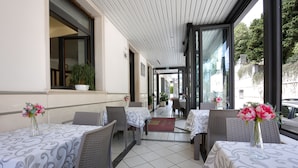 A covered outdoor dining area with tables and chairs, potted plants, and a view of the street. - Hotel Sollievo (San Giovanni Rotondo)