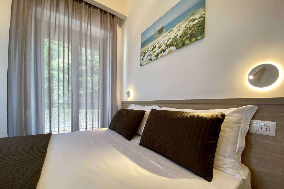Une chambre d’hôtel avec un grand lit, deux oreillers, une table de chevet, une lampe fixée au mur et une fenêtre avec des rideaux légers.