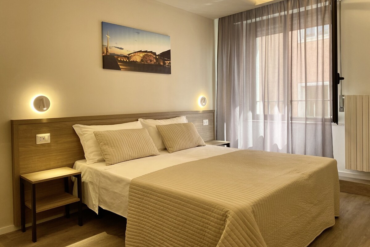 Une chambre d’hôtel avec un lit, des tables de chevet, un tableau et une fenêtre avec des rideaux.