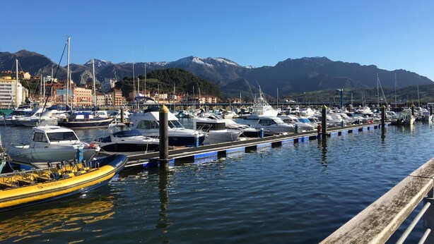 Un puerto deportivo con barcos amarrados, un muelle de madera y montañas al fondo.