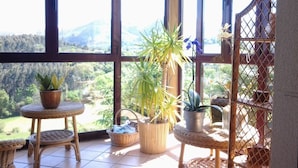 Un balcón con mobiliario de mimbre, plantas en macetas y vistas a un paisaje montañoso.