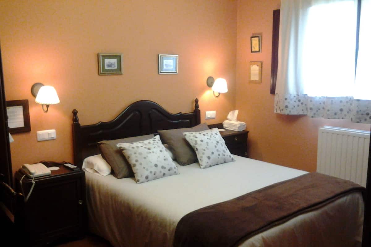 A neatly made bed with a brown headboard, two bedside lamps, a nightstand with a telephone, and a window with curtains.