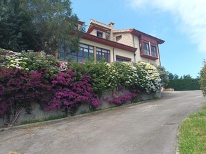 A two-story house with a red roof, large windows, and a garden with various flowers. - Hotel Foronda (Ribadesella)