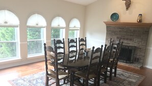 Comedor con una mesa redonda, seis sillas y un tapete estampado. Hay tres ventanas con cortinas blancas, una chimenea de ladrillo y un plato decorativo y un jarrón sobre la repisa.