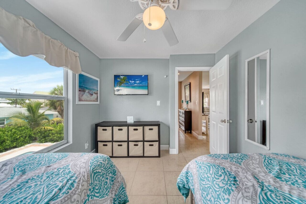 A bedroom with two beds, a ceiling fan, a TV, a desk with drawers, and a view of the ocean.