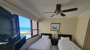 Een hotelkamer met twee bedden, een televisie, een plafondventilator, een schilderij en uitzicht op het strand.