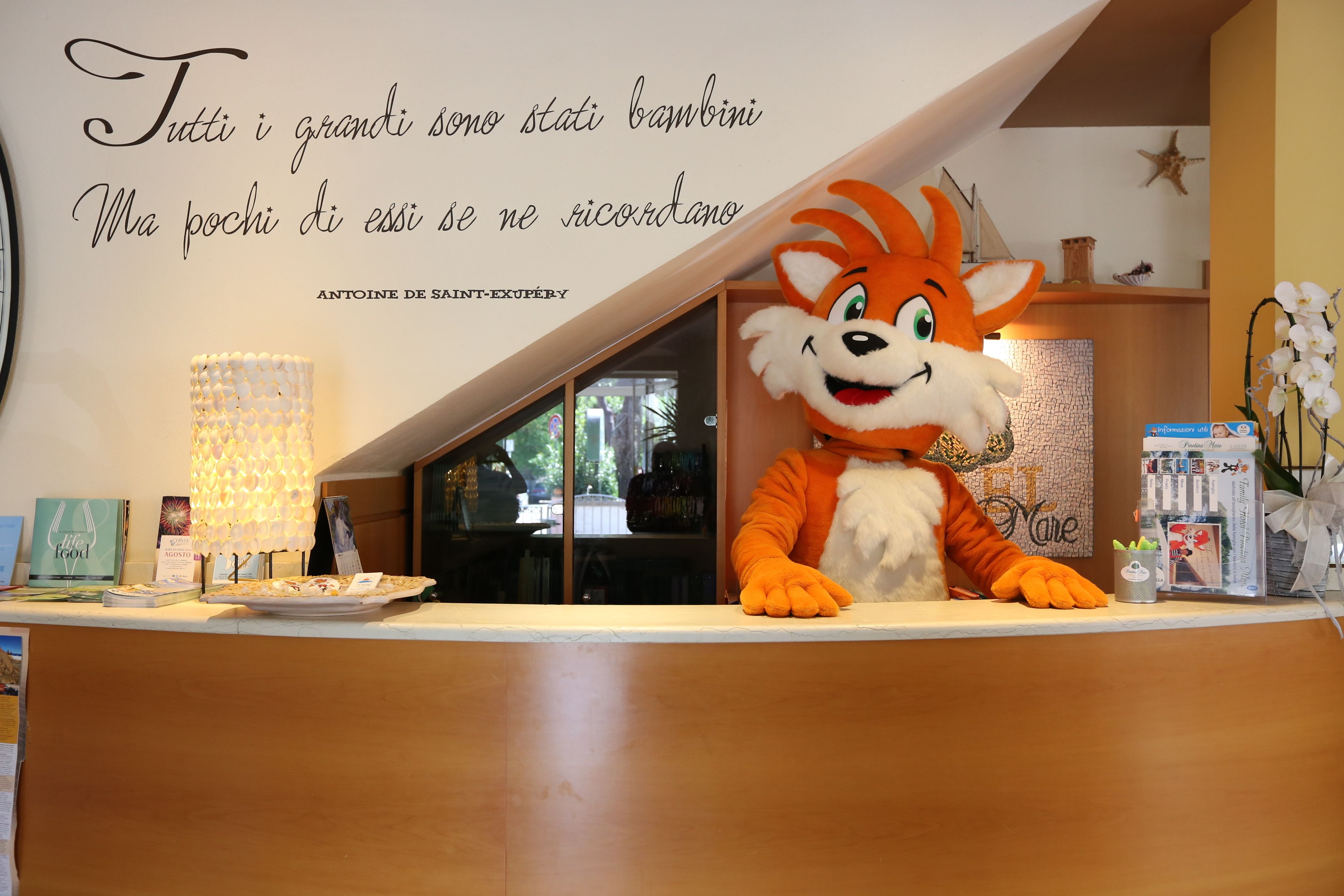 A hotel reception area with a large orange and white mascot, a quote on the wall, and a view of the interior through a glass partition.