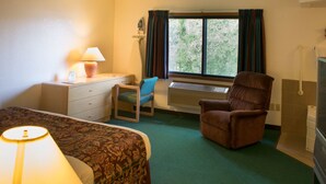 A hotel room with a bed, a desk with a lamp, a chair, a window with curtains, and a radiator. - Odisha House of Rest (Shawano)