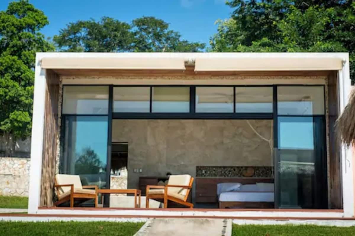 Un edificio moderno con un amplio frente de vidrio, un patio con mobiliario y un interior visible con una cama y chimenea.