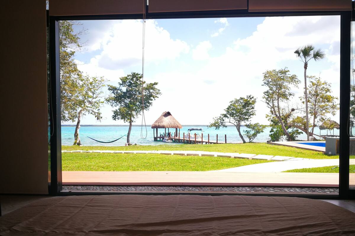 Una habitación con un ventanal grande que da a una playa, una construcción de techo de paja y una hamaca.