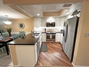 A kitchen with white cabinets, stainless steel appliances, a wooden floor, and a dining area with a clock on the wall. - Sea Winds 30 Ocean View Retreat (St. Augustine)