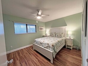 A bedroom with a bed, a ceiling fan, a window with blinds, and a nightstand with a lamp. - Sea Winds 30 Ocean View Retreat (St. Augustine)