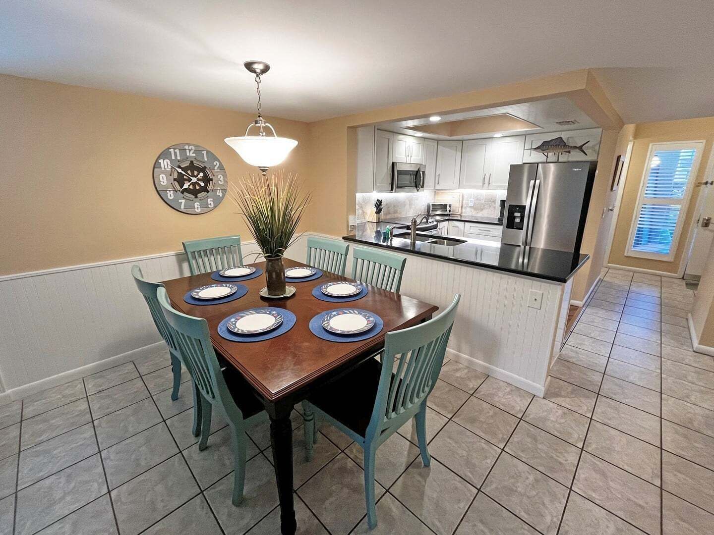 A dining area with a wooden table set for six, surrounded by teal chairs, a wall clock, and a hanging light fixture.