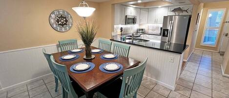 A dining area with a wooden table set for six, surrounded by teal chairs, a wall clock, and a hanging light fixture.