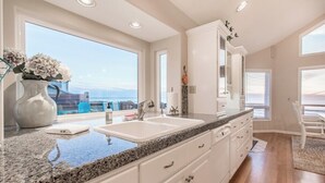 A modern kitchen with white cabinets, granite countertops, and a large window offering a view of the ocean. - Top O' the Dune · Luxury Oceanfront, Huge home & views, game room (Pacific City)