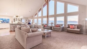 A spacious living room with large windows, a kitchen in the background, and a view of the sea. - Top O' the Dune · Luxury Oceanfront, Huge home & views, game room (Pacific City)