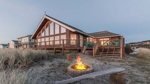 A wooden cabin with a fire pit in the foreground, a staircase leading to a deck, and a view of the surrounding landscape. - Top O' the Dune · Luxury Oceanfront, Huge home & views, game room (Pacific City)