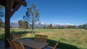 A wooden deck with a table and chairs, overlooking a grassy field and mountains.