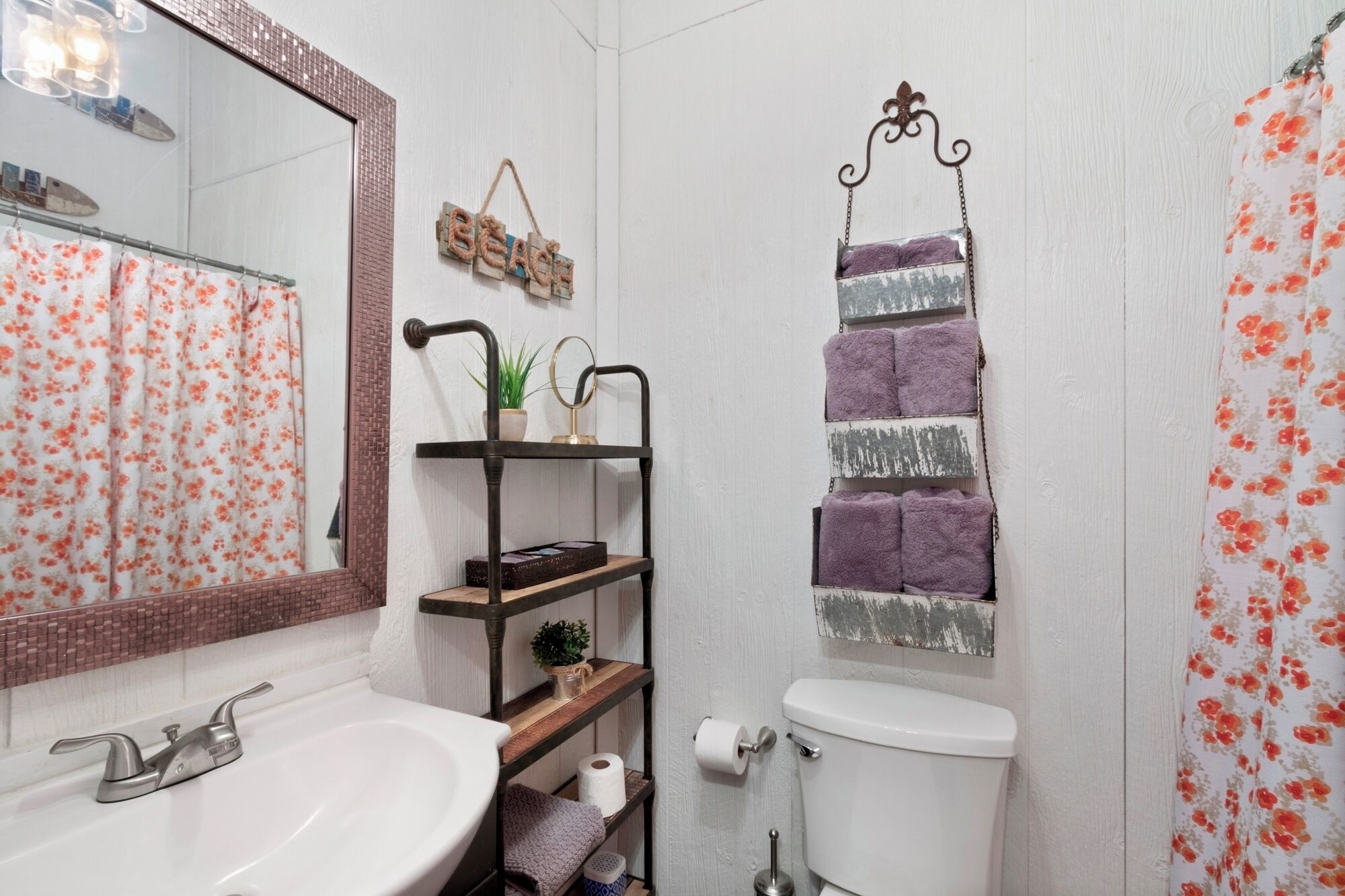 A bathroom with a white sink, a toilet, a mirror, a shelf with plants, and a towel rack with purple towels.