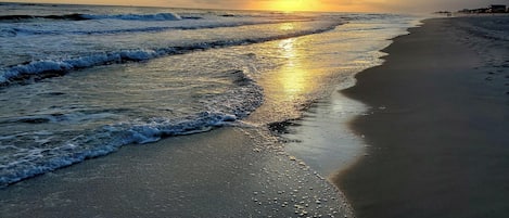 Une plage avec des vagues, un coucher de soleil et une côte lointaine parsemée de bâtiments.