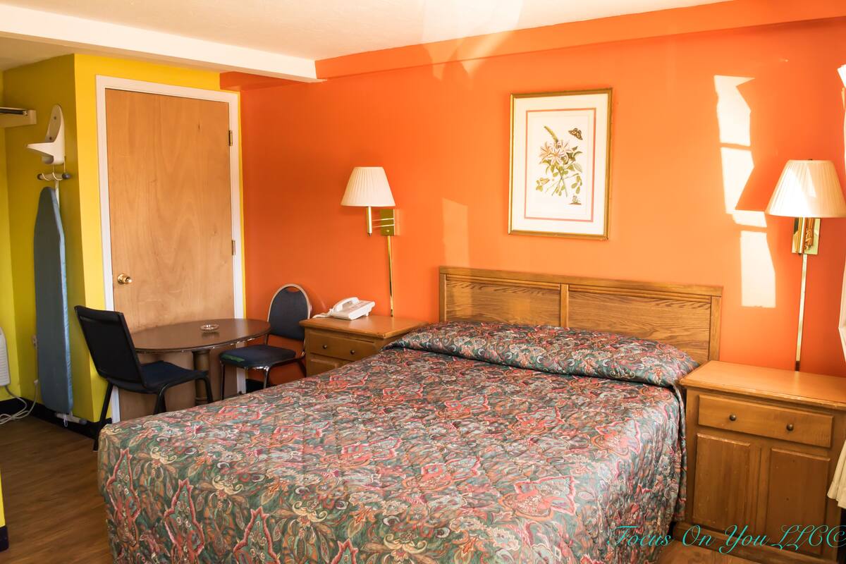 A hotel room with a bed, a desk, a chair, a lamp, and a door.