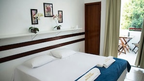 Habitación de hotel con cama, paredes blancas, cabecera de madera, un pequeño estante con cuadros enmarcados y una puerta a un balcón.