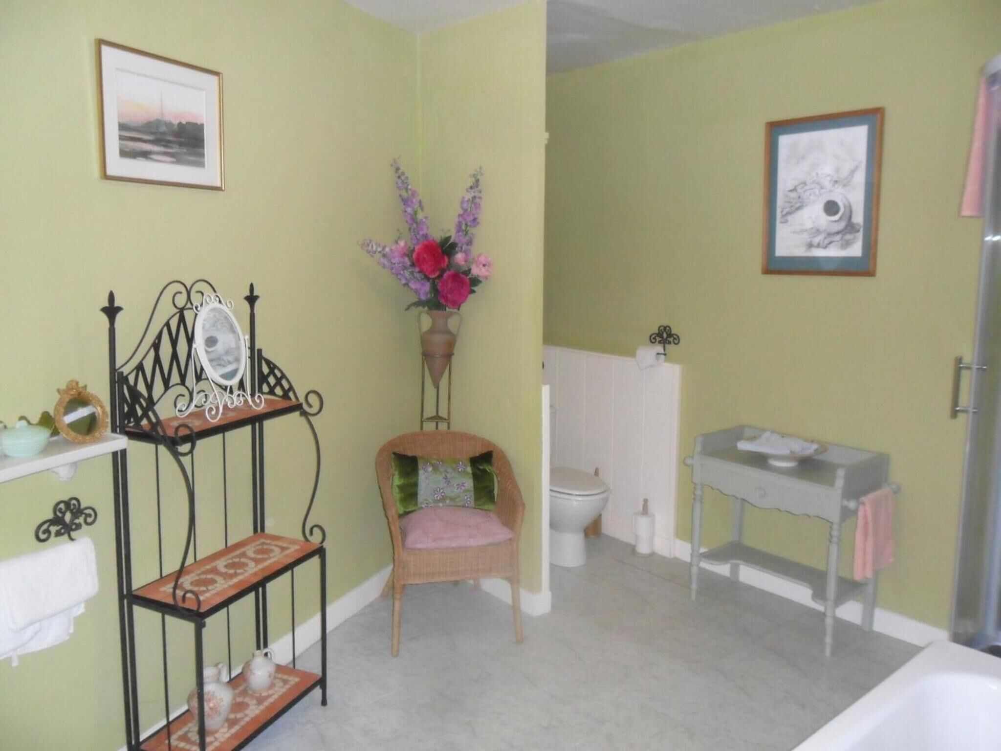 A bathroom with a sink, toilet, and chair. There is a mirror on a stand, a vase with flowers, and a framed picture on the wall.