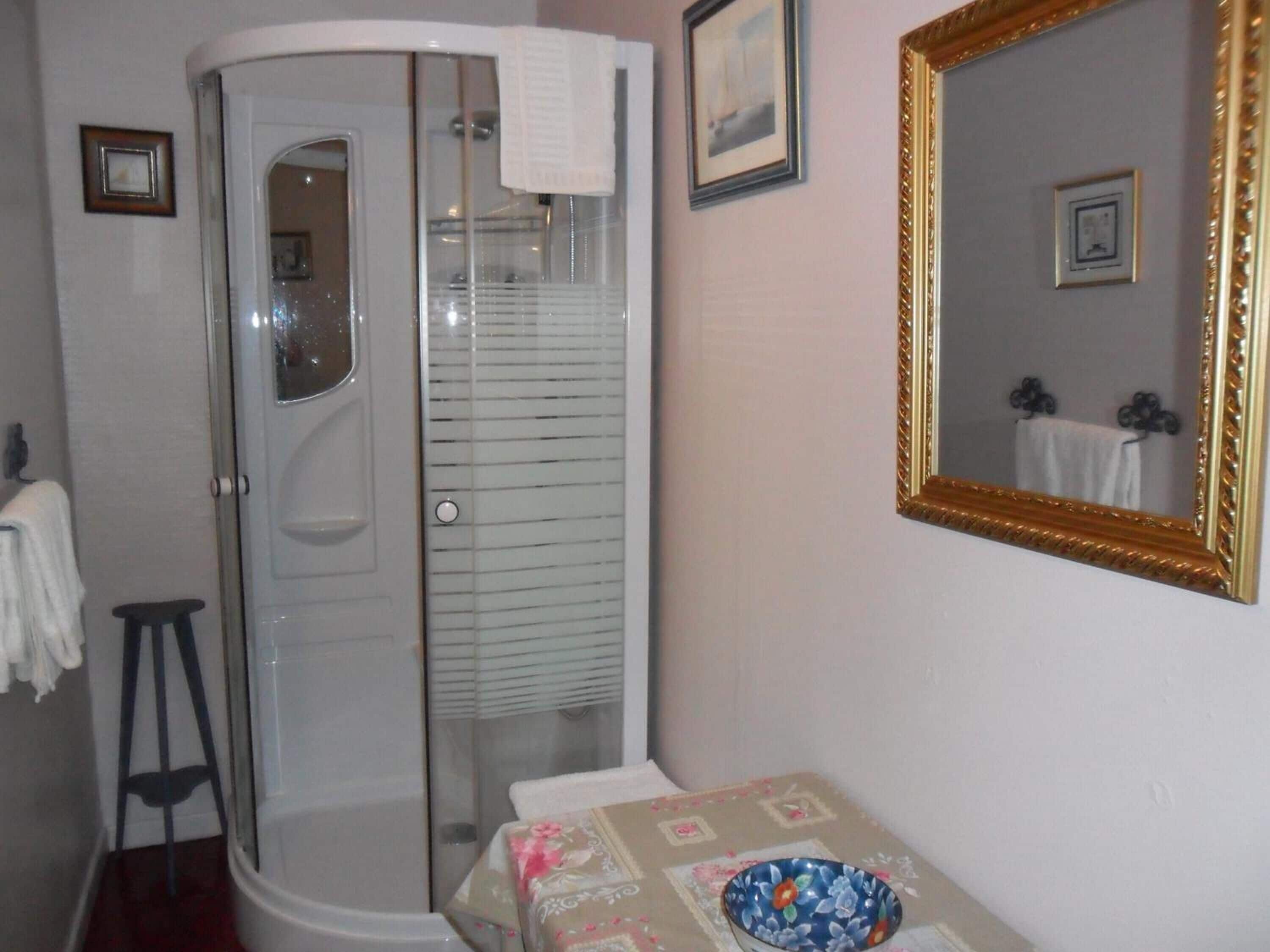 A bathroom with a glass-enclosed shower, a sink, a mirror with a decorative frame, and a small stool.