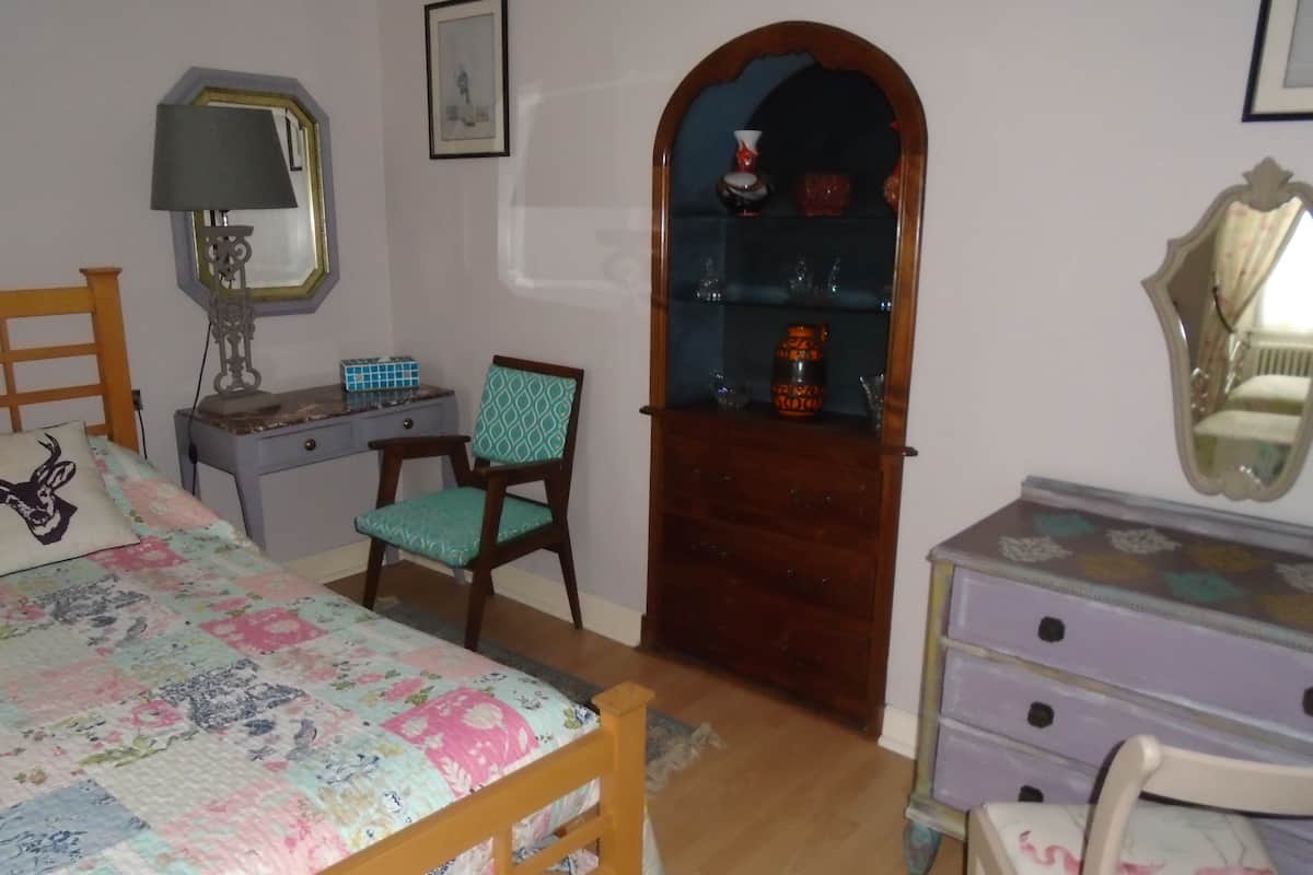 Ein Schlafzimmer mit einem Bett, einem Sessel, einem Schminktisch, einem Spiegel und einem Regal mit Dekoration.