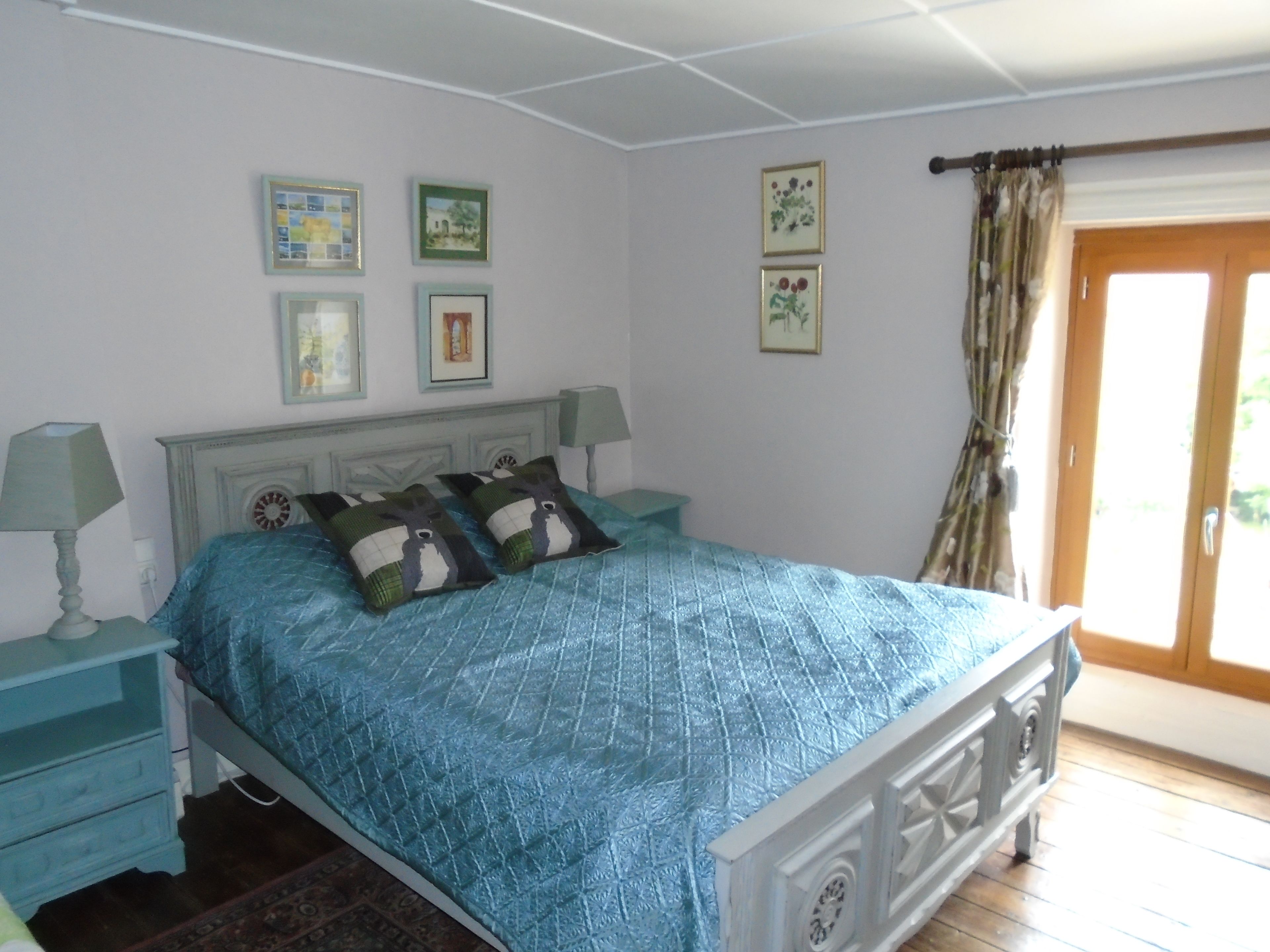 Un dormitorio con una cama, dos mesitas de noche, una ventana con cortinas y cuadros en la pared.