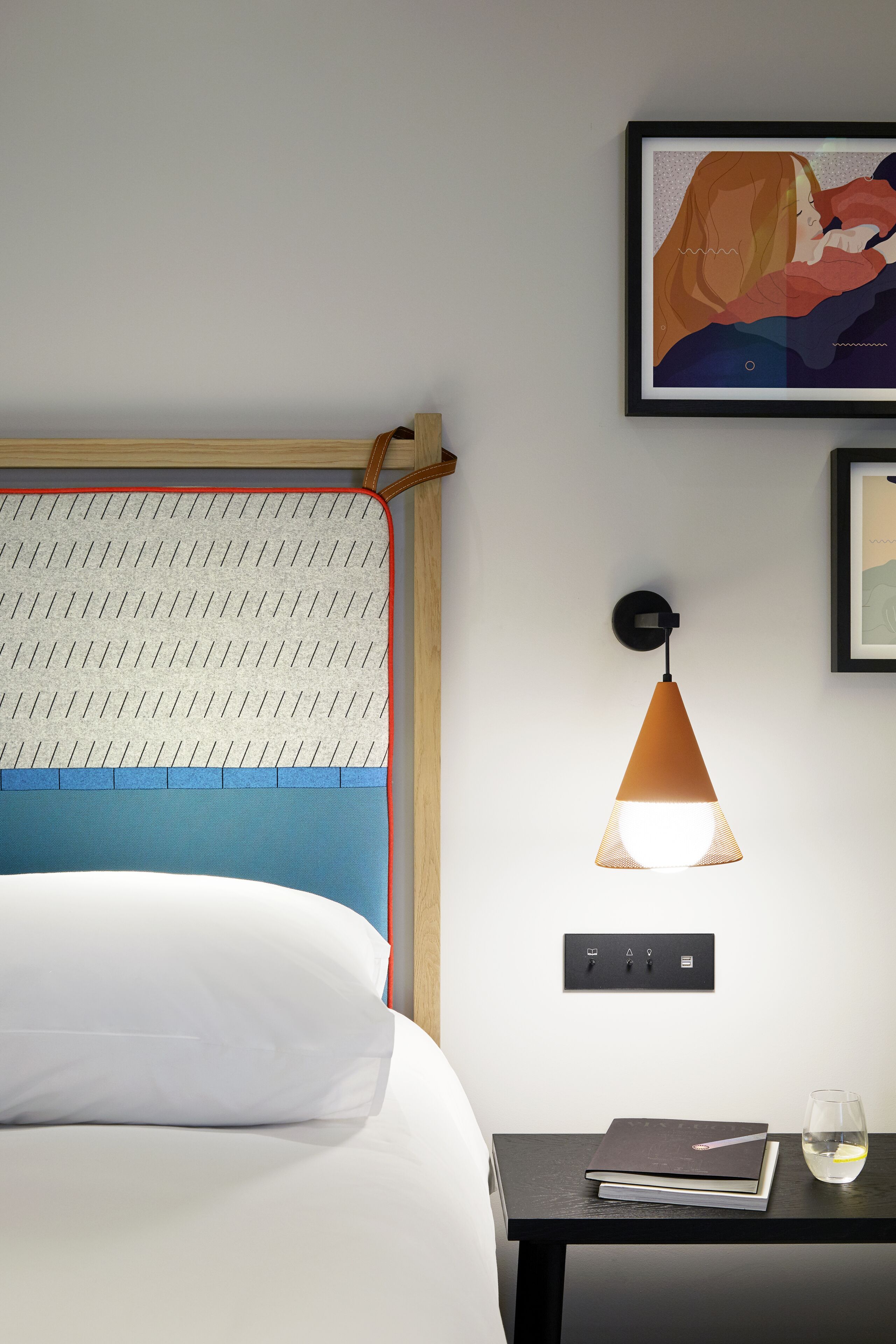 A neatly made bed with a headboard, a bedside table with a book and a glass of water, and a wall-mounted lamp.