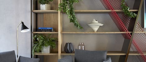 Un salon moderne avec un canapé gris, un lampadaire noir et une bibliothèque agrémentée de plantes et d’objets décoratifs.