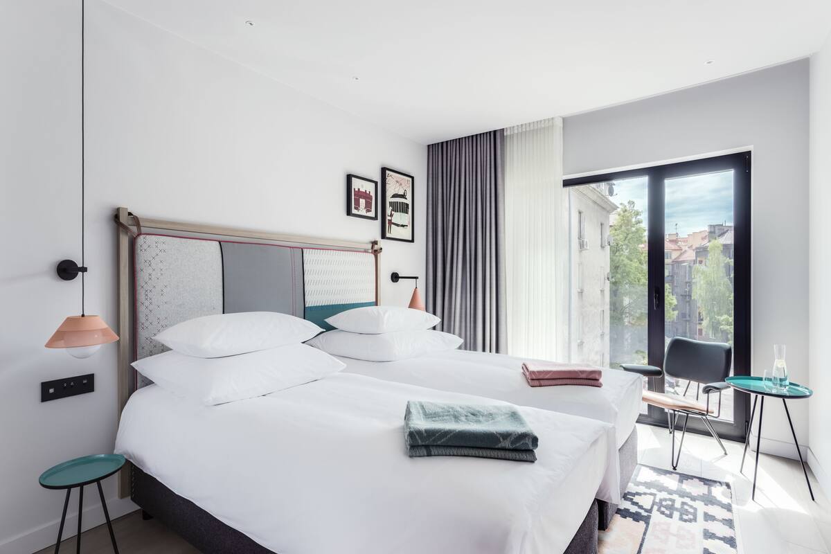 A modern hotel room with a large bed, a small table, a chair, and a view of the city through the window.