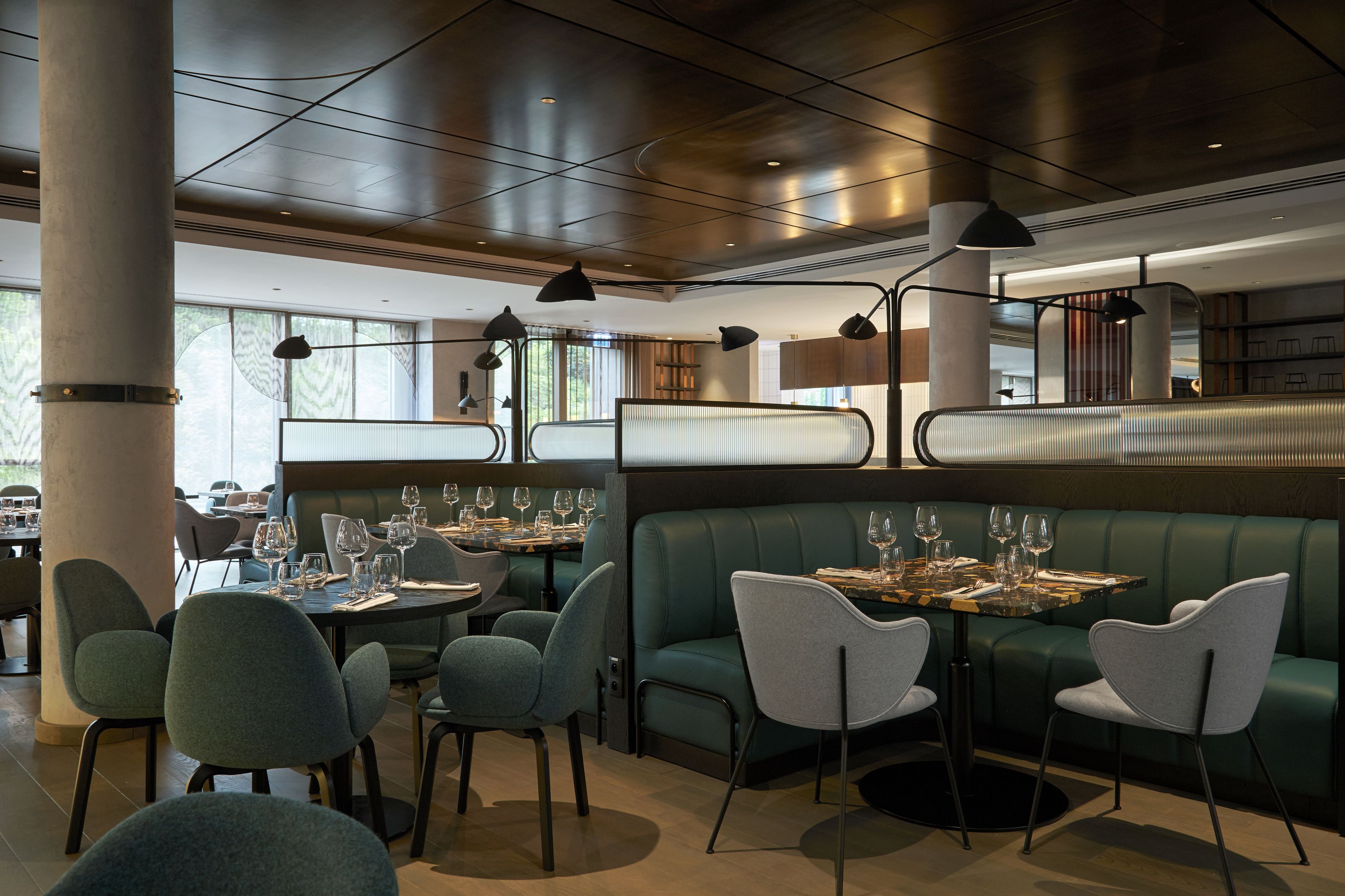 Un restaurante moderno con asientos tipo banqueta verdes y mesas redondas, con iluminación colgante y vistas al exterior.