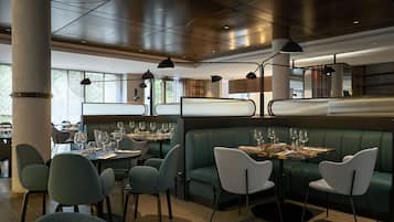 Un restaurante moderno con asientos tipo banqueta verdes y mesas redondas, con iluminación colgante y vistas al exterior.