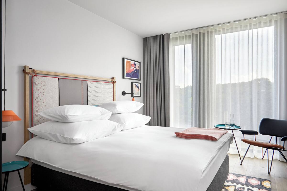 A hotel room with a bed, a chair, a small table, and a window with curtains.