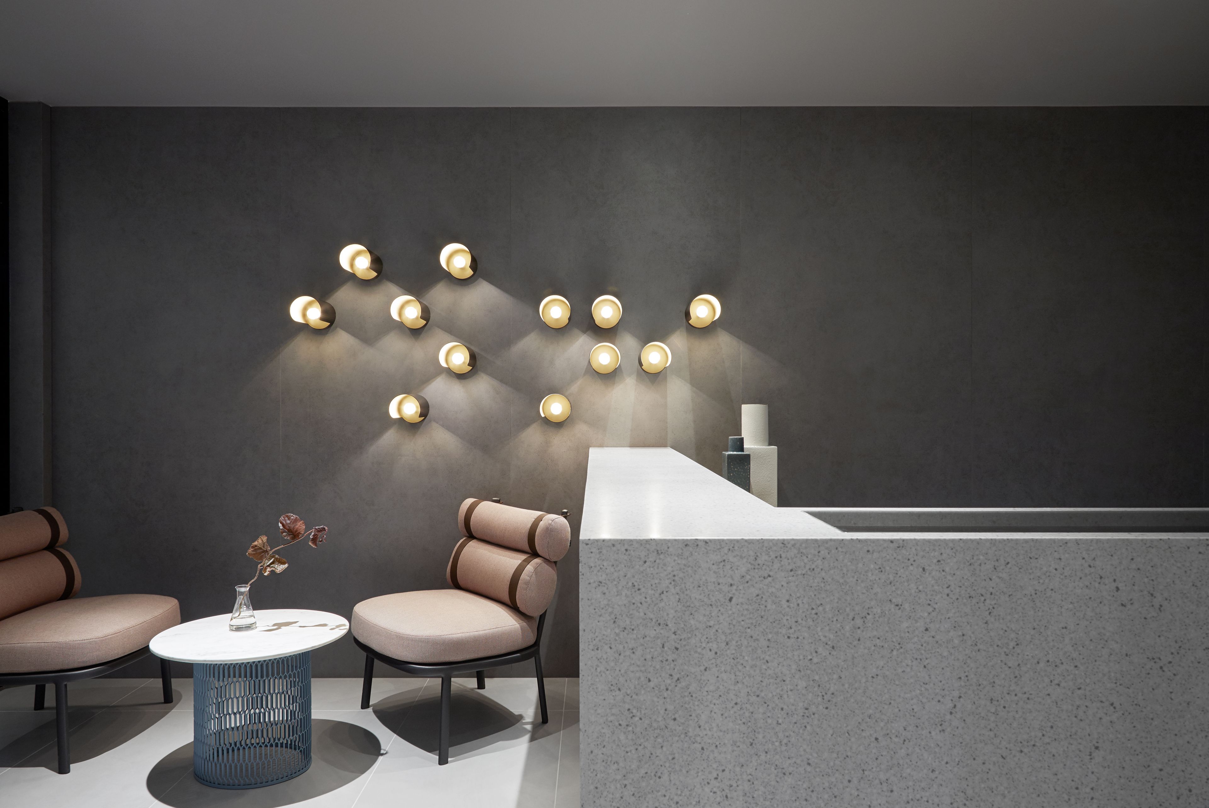 Un espace d’accueil moderne avec un comptoir de réception blanc, deux fauteuils confortables, une petite table bleue et un luminaire fixé au mur.