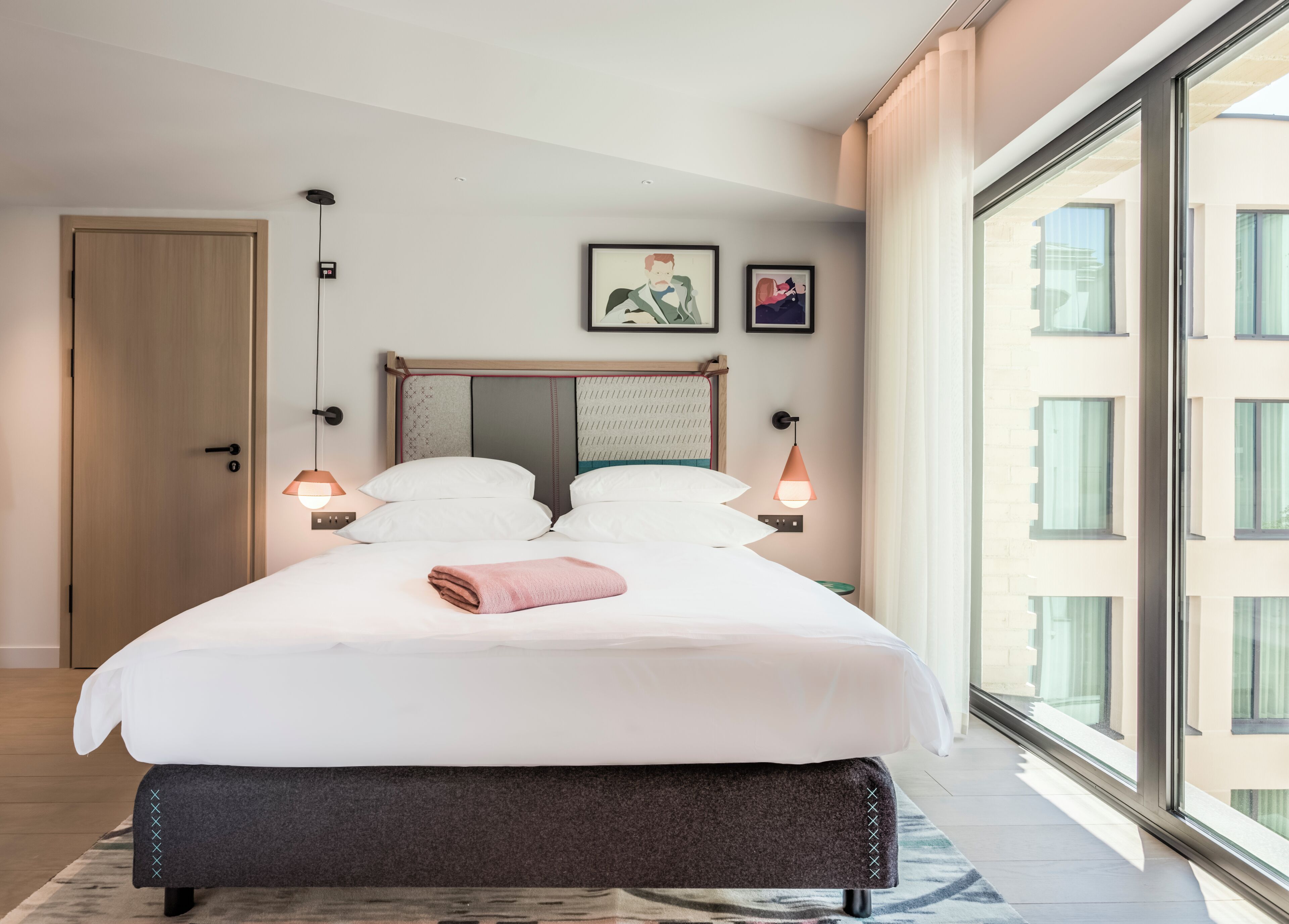 Habitación de hotel moderna con una cama grande, vistas a través de ventanales del suelo al techo y una puerta a la izquierda.