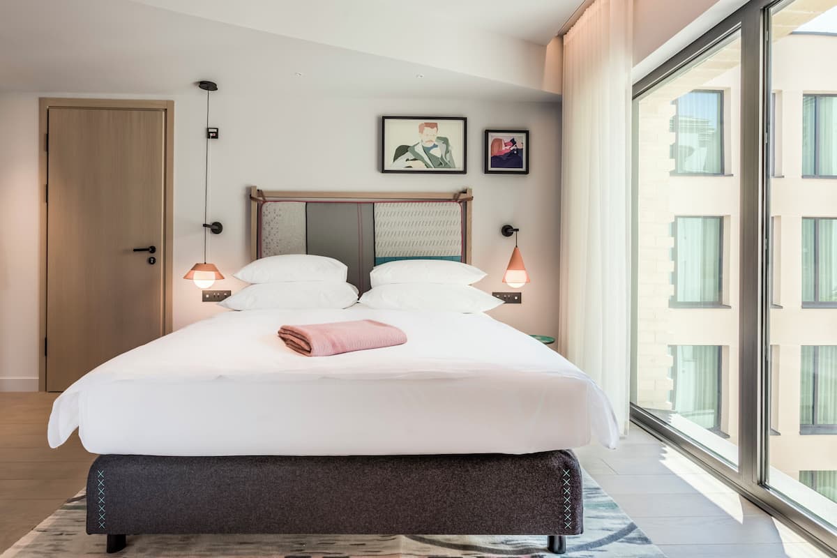 A modern hotel room with a large bed, a view through floor-to-ceiling windows, and a door to the left.