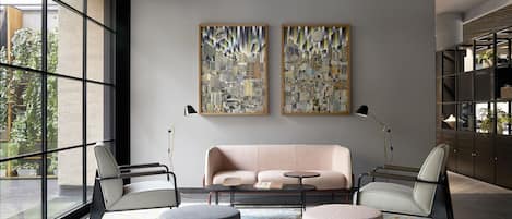 A modern living room with a sofa, two armchairs, a coffee table, and a patterned rug. There are two large paintings on the wall and a large window with a view of greenery outside.